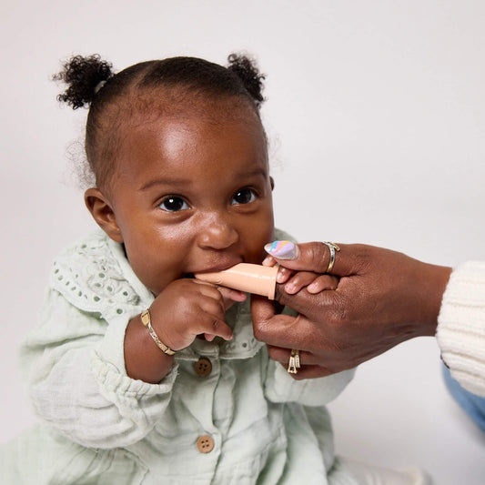 A baby in a pale green dress bites a peach silicone finger toothbrush while a woman gently holds it in place.