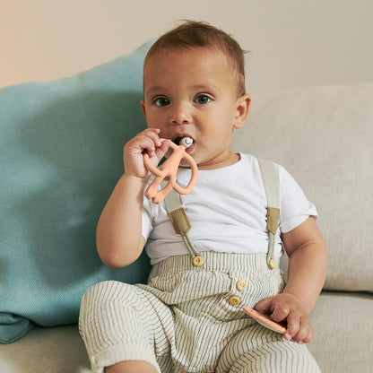 A toddler sitting on a sofa chews a peach animal-shaped silicone teether while holding a matching flat teether.