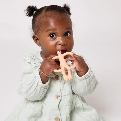 A baby in a pale green dress holds and chews a peach animal-shaped silicone teether with easy-grip handles.