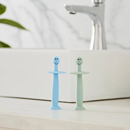 Two silicone baby training toothbrushes standing upright on a bathroom counter beside a sink with suction bases attached.