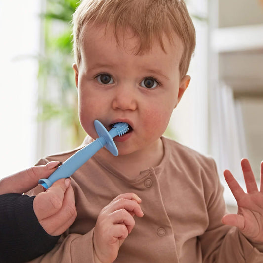 Baby holding a silicone training toothbrush with wide safety guard while an adult hand guides brushing near the mouth.