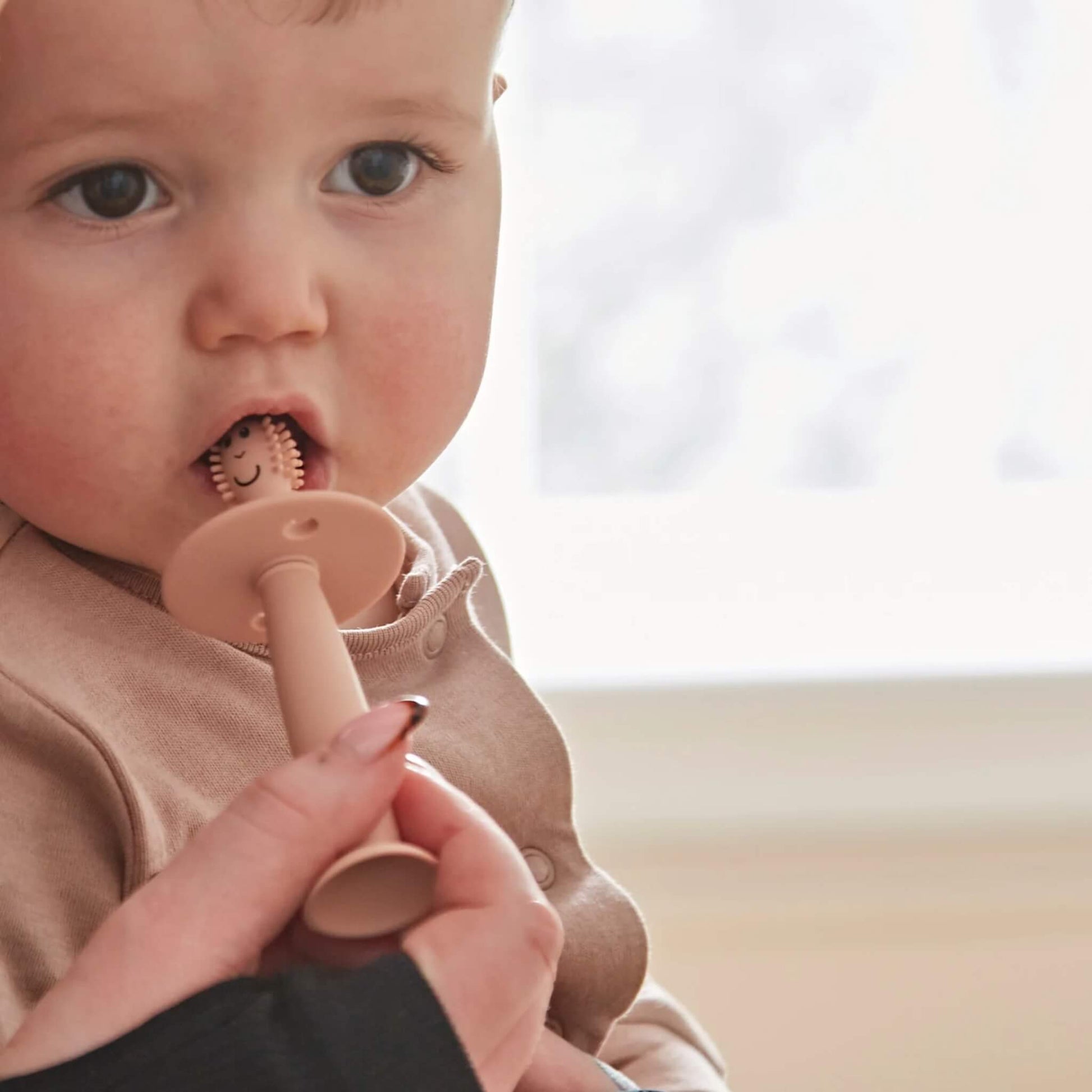 Baby holding a silicone training toothbrush with wide safety guard while an adult hand guides brushing near the mouth.