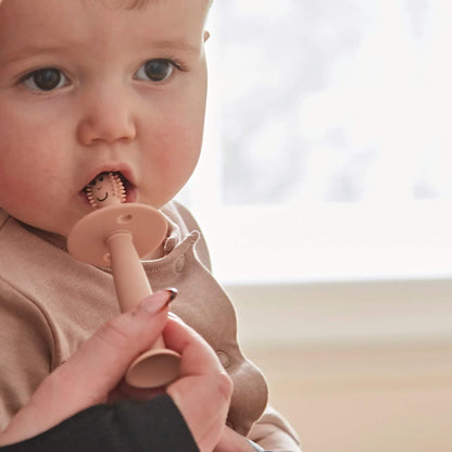 Baby holding a silicone training toothbrush with wide safety guard while an adult hand guides brushing near the mouth.