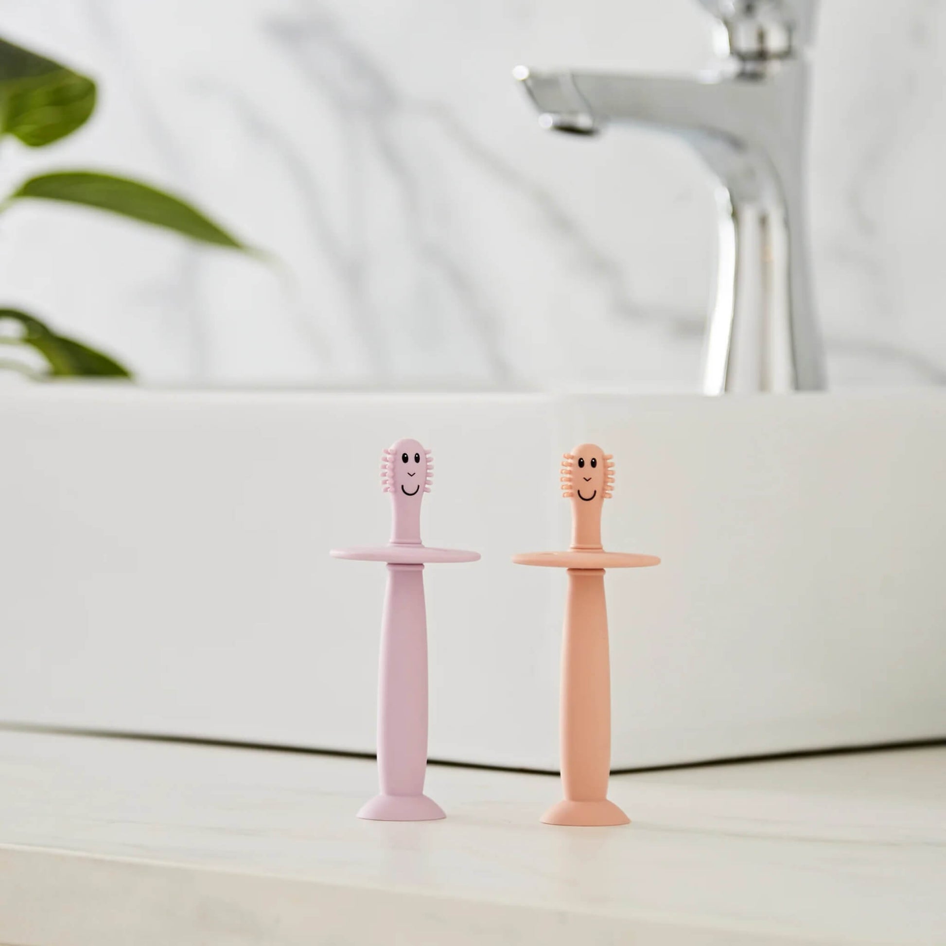 Two silicone baby training toothbrushes standing upright on a bathroom counter beside a sink with suction bases attached.