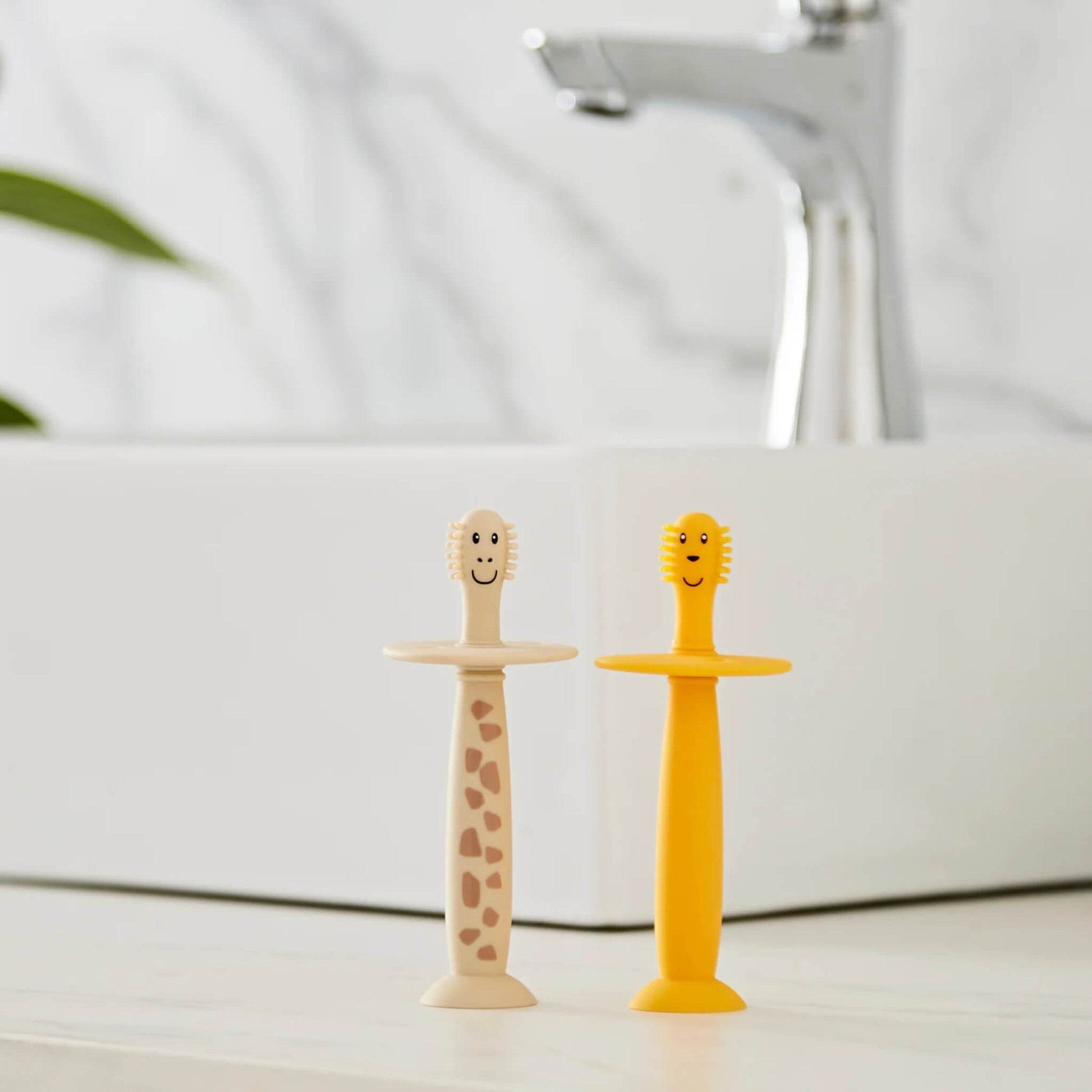 Two silicone baby training toothbrushes standing upright on a bathroom counter beside a sink with suction bases attached