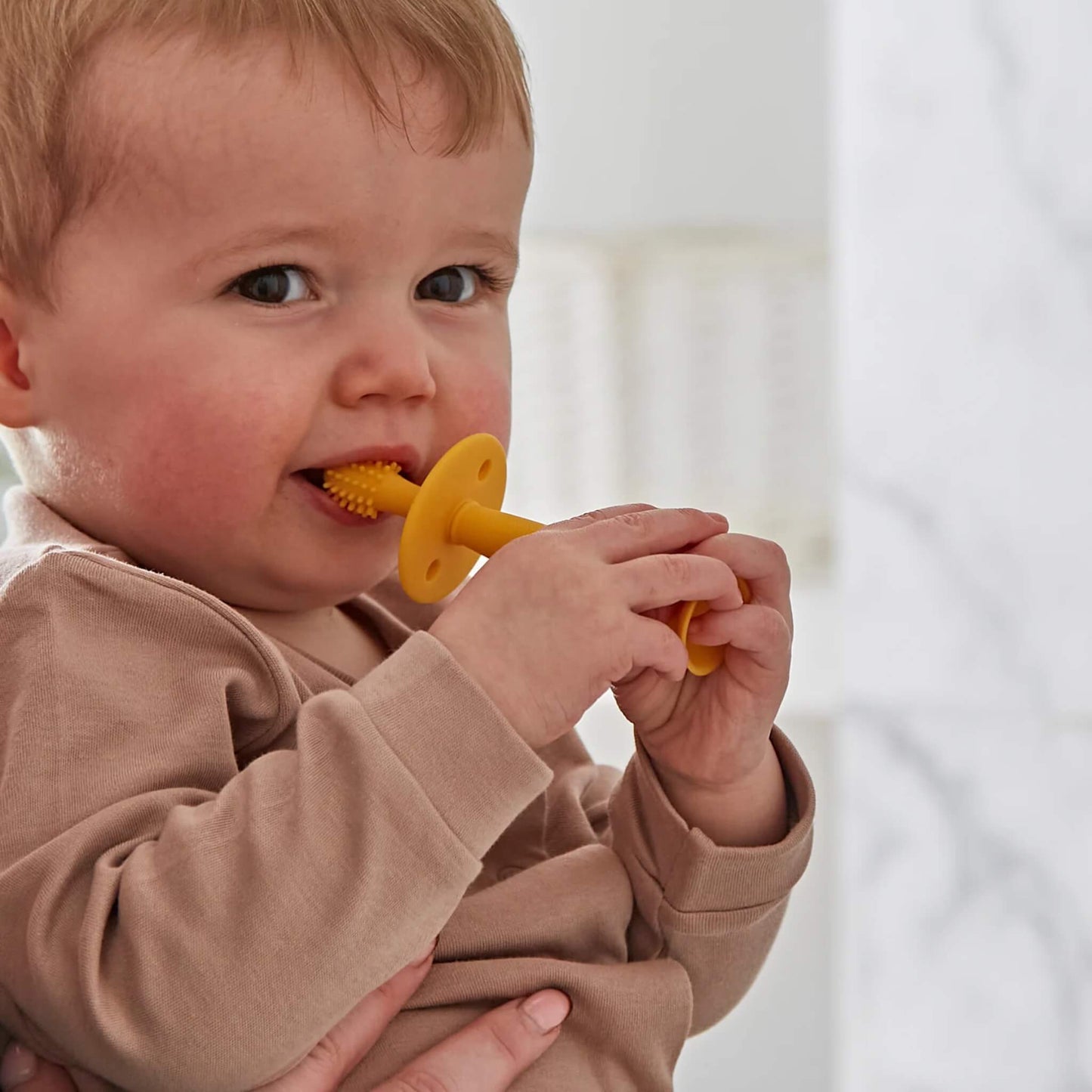 Baby holding a silicone training toothbrush with wide safety guard while an adult hand guides brushing near the mouth.