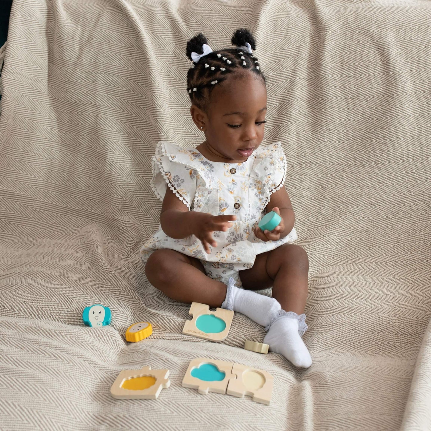 A toddler sitting on a patterned blanket holding one of the puzzle pieces while the rest of the wooden animal set is spread out nearby.