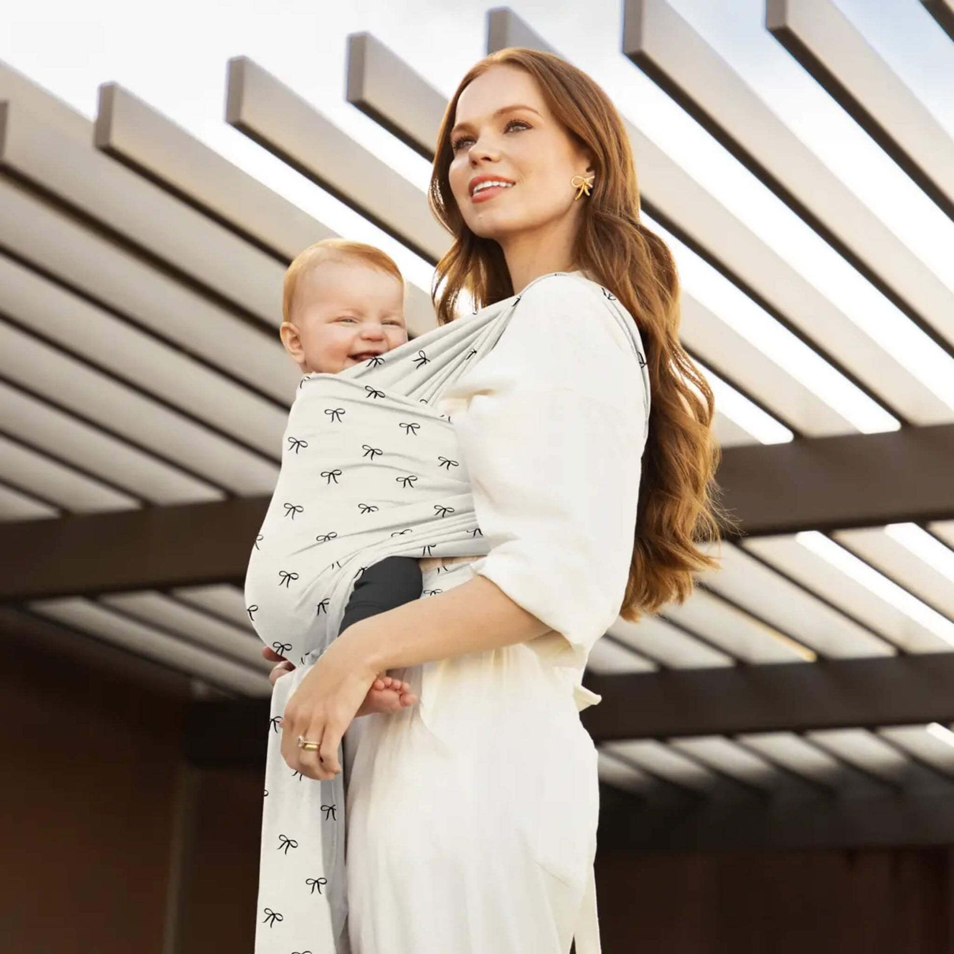 Parent standing outdoors carrying a baby in a light wrap patterned with small bows, with the wrap tied at the side and the baby resting against the chest.