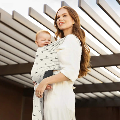 Parent standing outdoors carrying a baby in a light wrap patterned with small bows, with the wrap tied at the side and the baby resting against the chest.