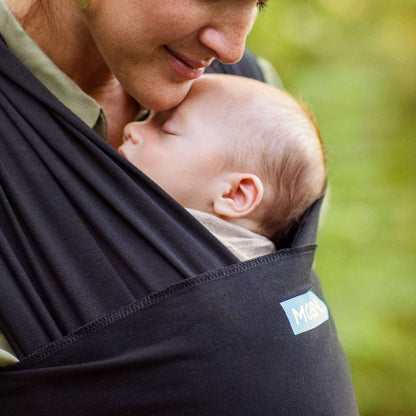 Mother gently kissing her baby’s head while they rest in a soft black wrap carrier.