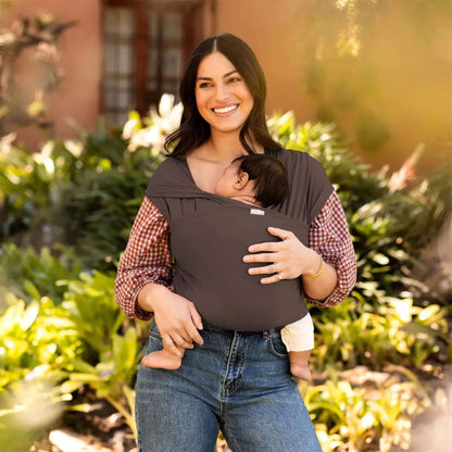 Mother smiling outdoors while carrying her baby in a Hickory brown baby wrap carrier surrounded by greenery.