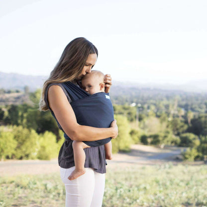 Mother standing outdoors kissing her baby’s head while carrying them in a navy baby wrap carrier.