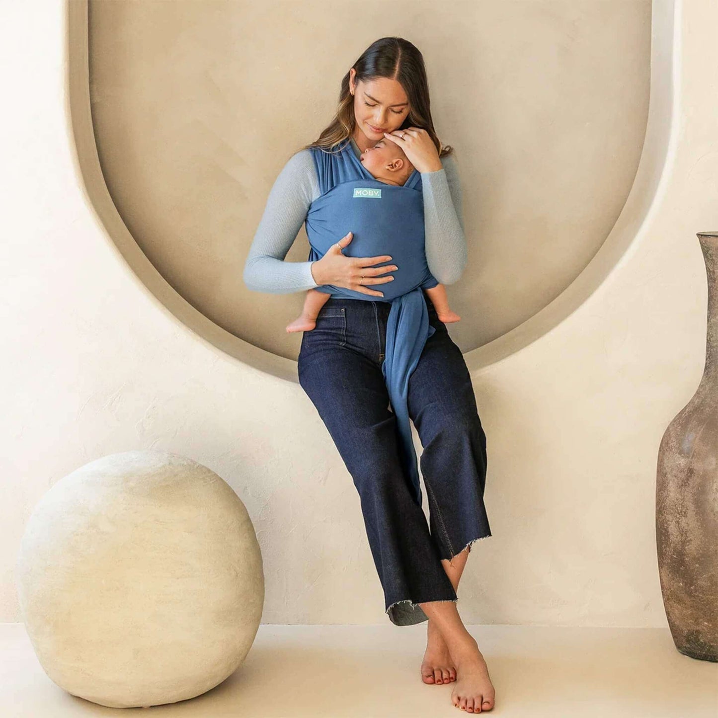 Mother sitting indoors with her baby resting in a soft Ocean blue wrap carrier, gently touching their head.