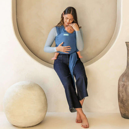 Mother sitting indoors with her baby resting in a soft Ocean blue wrap carrier, gently touching their head.