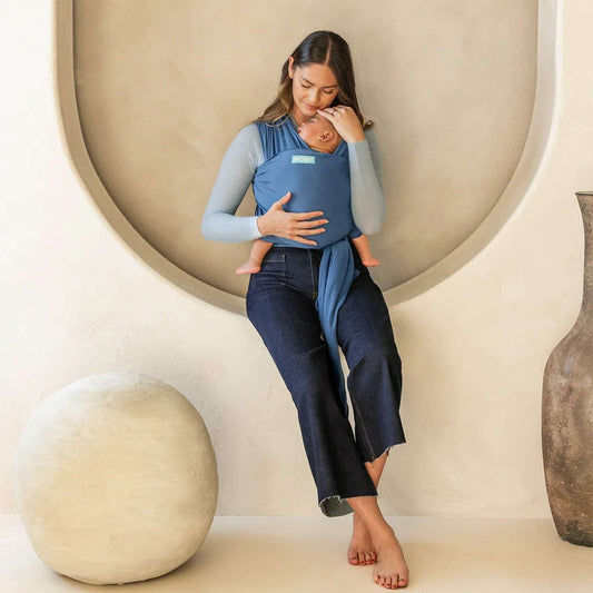 Mother sitting indoors with her baby resting in a soft Ocean blue wrap carrier, gently touching their head.