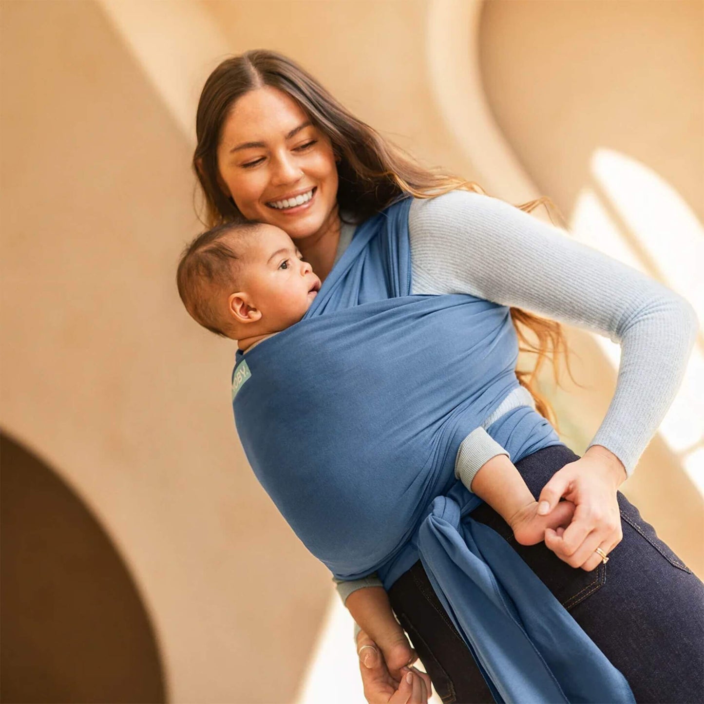 Mother smiling while carrying her baby in an Ocean blue wrap carrier indoors with soft natural light.