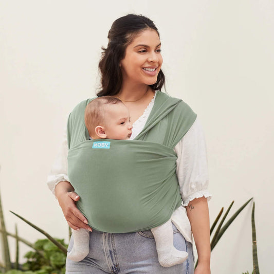 Mother smiling while carrying her baby in a Pear green wrap carrier indoors with light plants in the background.