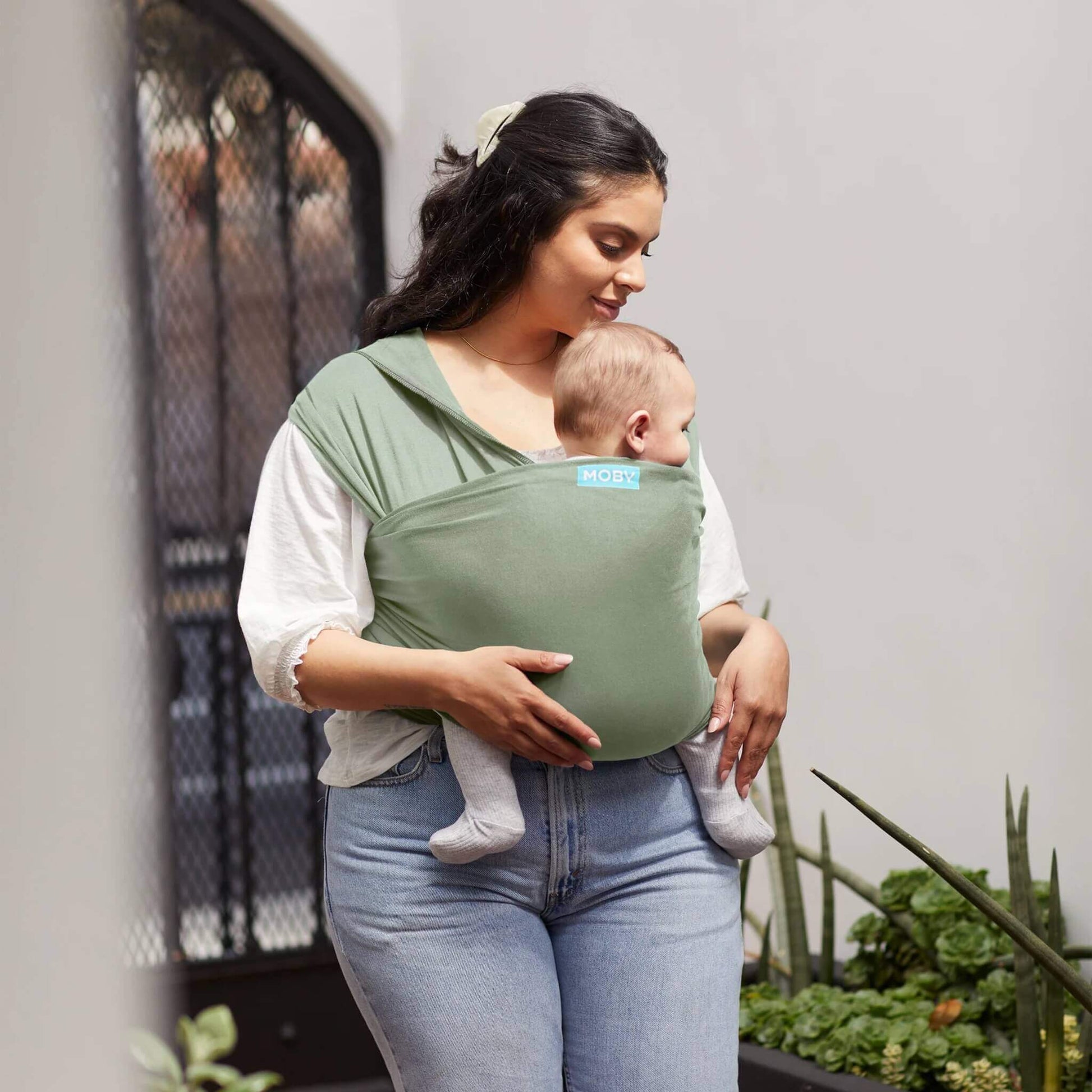Mother standing near plants holding her baby in a Pear green wrap carrier while gently looking down at them.