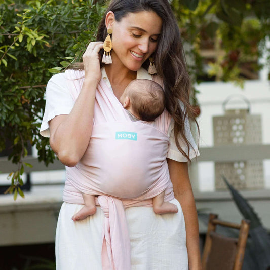 Mother smiling while cradling her baby in a Rose Quartz pink wrap carrier outdoors surrounded by greenery.