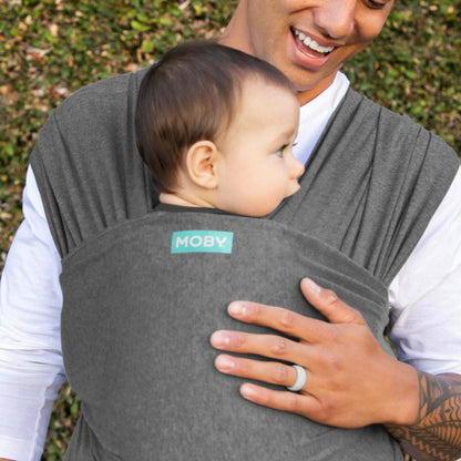 Adult holding a baby in a deep grey wrap outdoors, with one hand supporting the baby and the wrap covering the baby’s back.