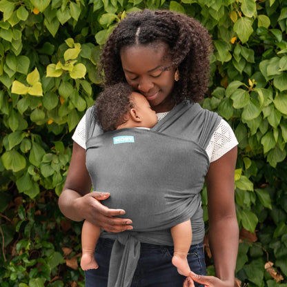 Parent standing in a deep grey wrap while holding a sleeping baby close against their chest, with the wrap tied securely at the waist.