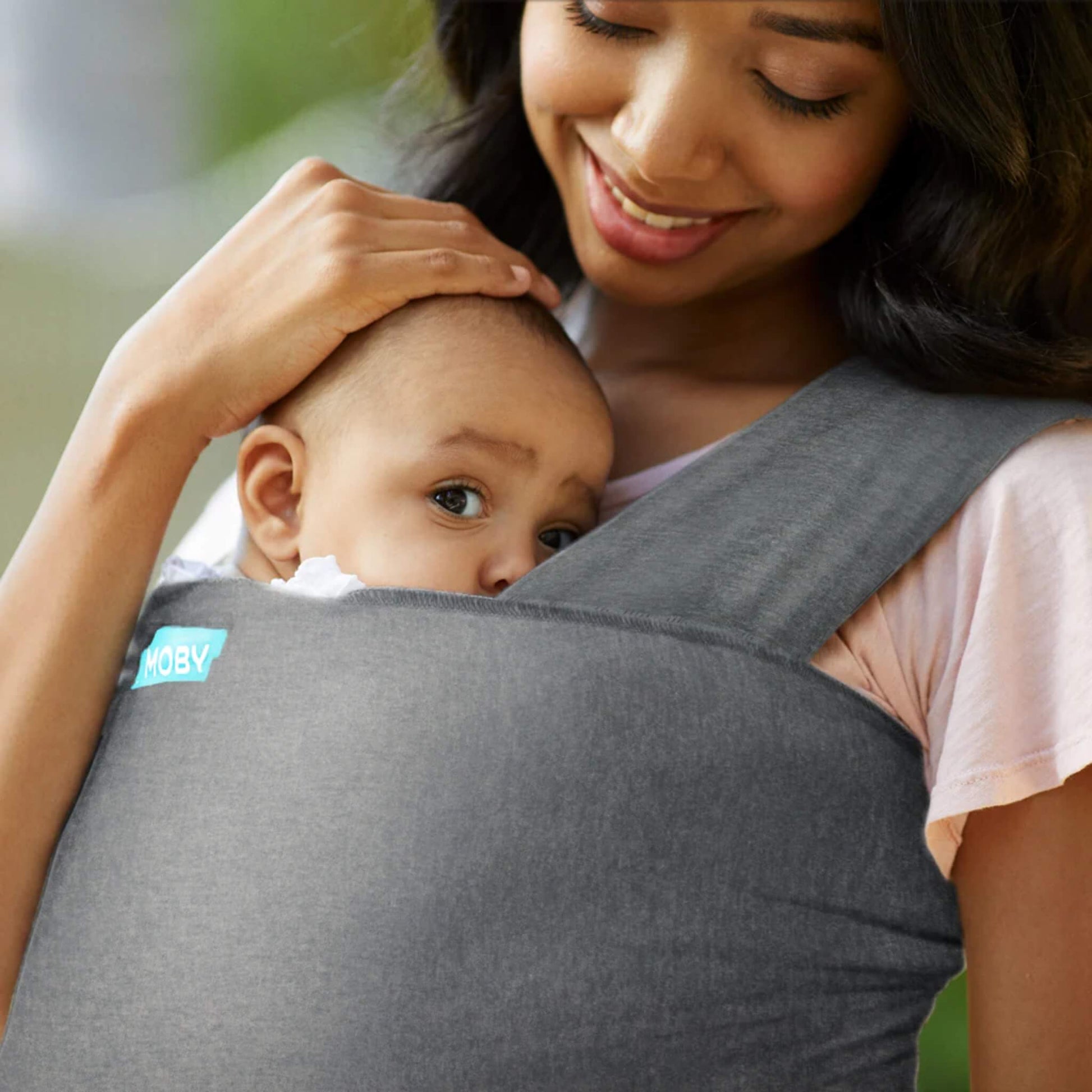Baby resting against an adult’s chest in a deep grey wrap, with the adult’s hand gently placed on the baby’s head.