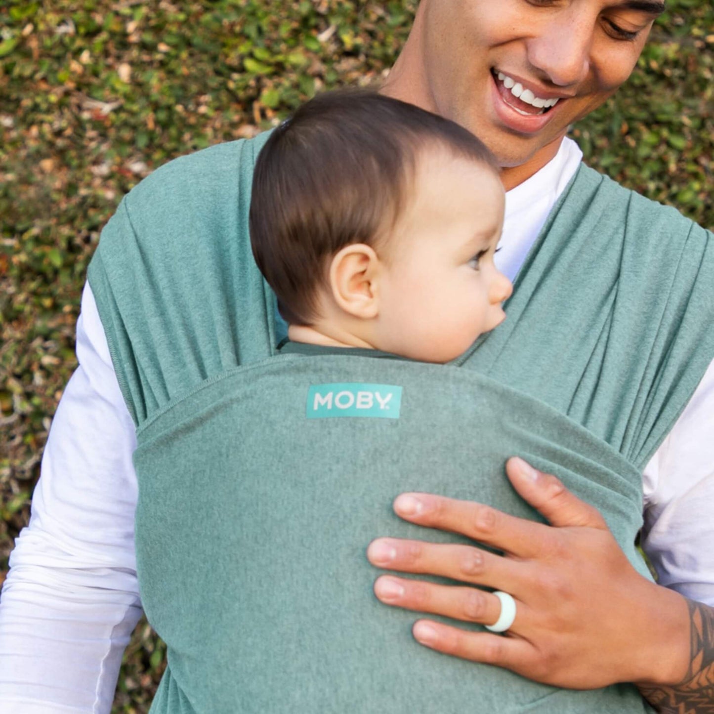 Baby resting against an adult’s chest while being carried in a teal wrap, with the baby’s face turned outward and the adult smiling down.