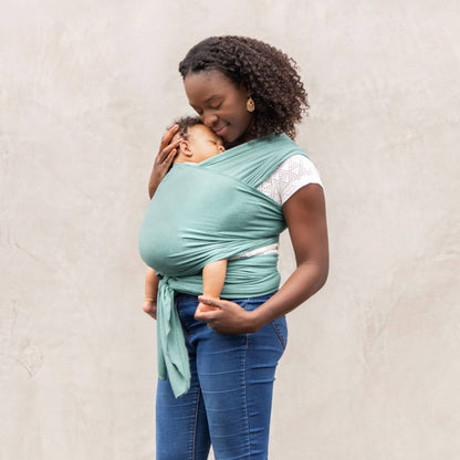 Parent walking outdoors while holding a baby in a teal wrap, with the baby facing inward and the adult supporting them with one hand.