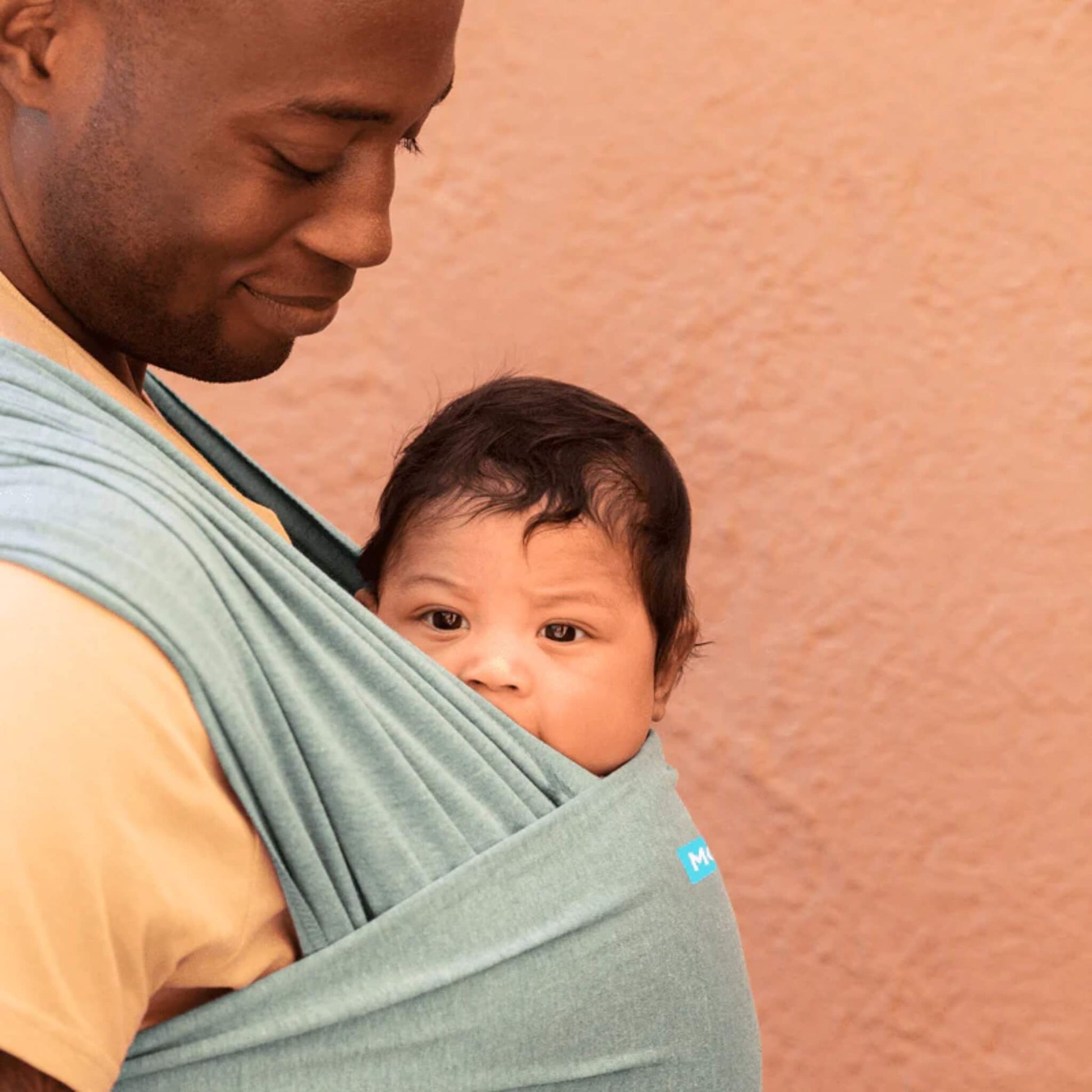 Baby being carried close against an adult’s chest inside a teal wrap, with the baby looking out from the top edge of the fabric.