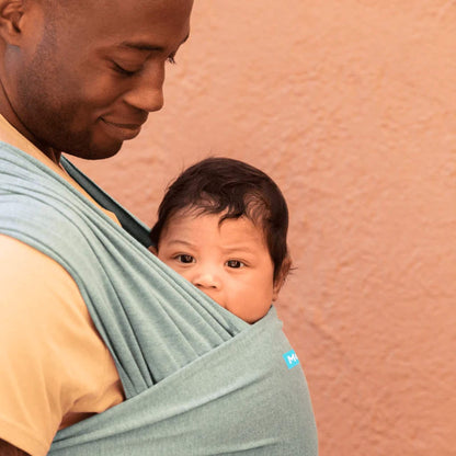 Baby being carried close against an adult’s chest inside a teal wrap, with the baby looking out from the top edge of the fabric.