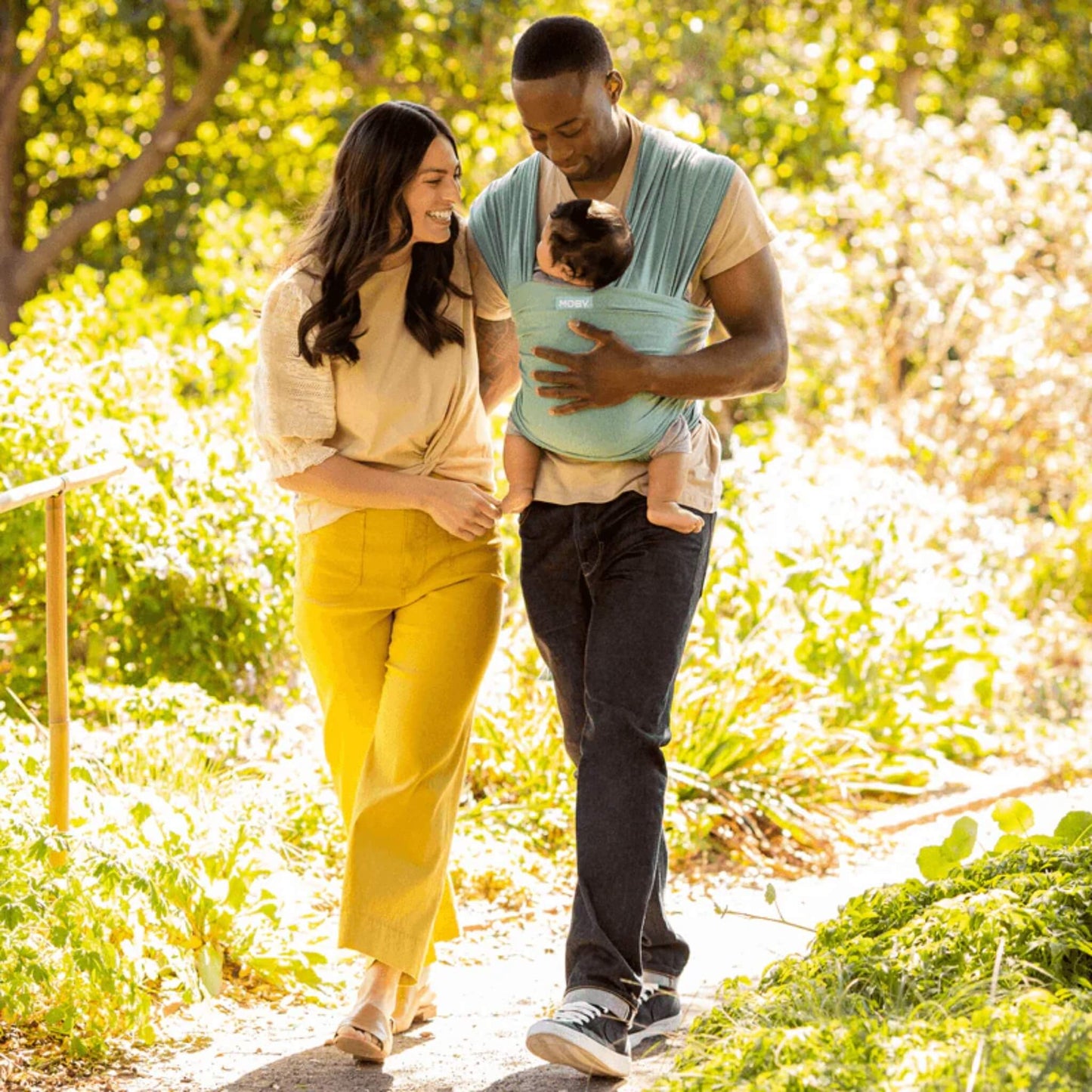 Two adults walking outdoors, with one carrying a baby in a teal wrap while the other walks beside them, surrounded by greenery.