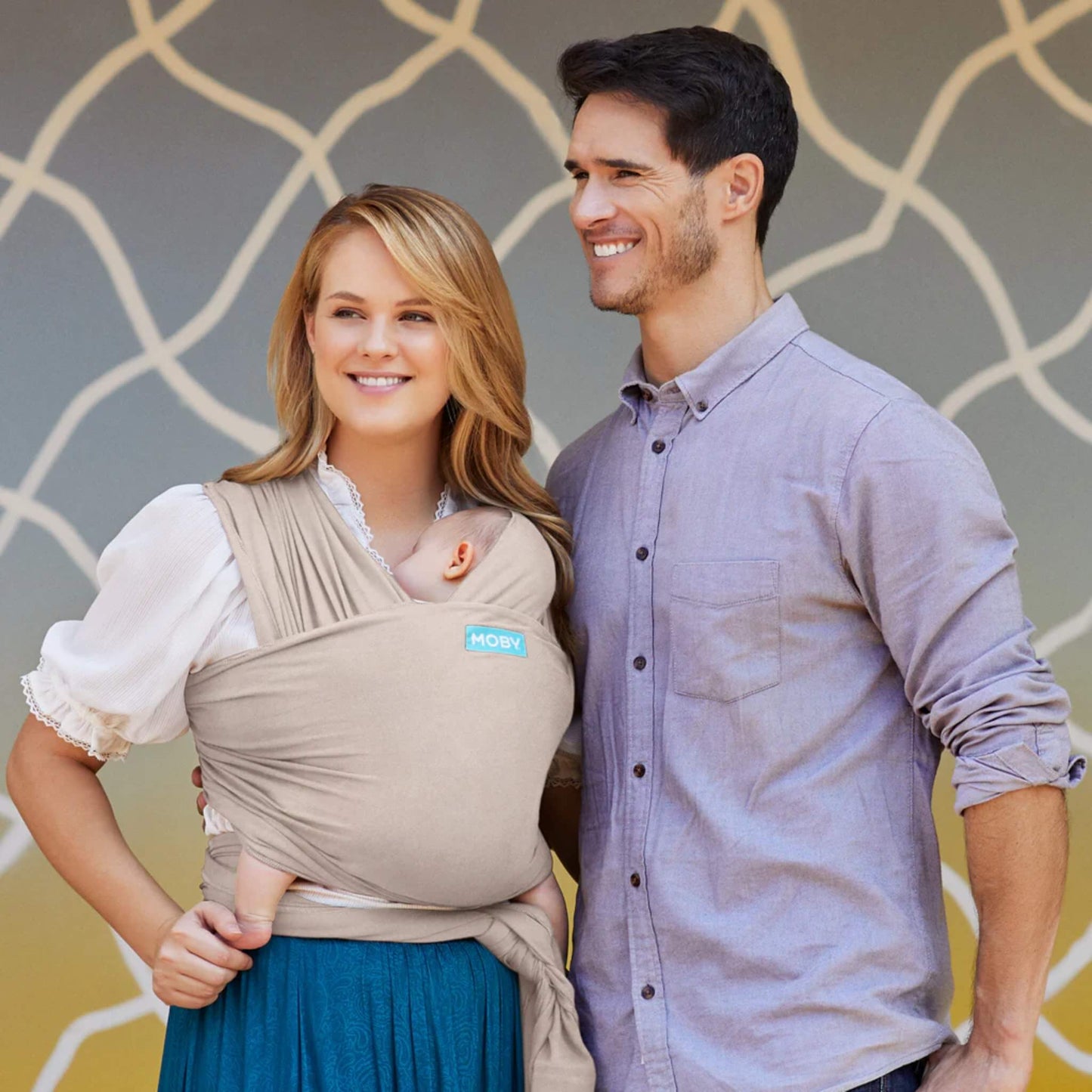 Parent holding a baby in a front-carry wrap while standing beside a partner, with the wrap supporting the baby close against the chest.