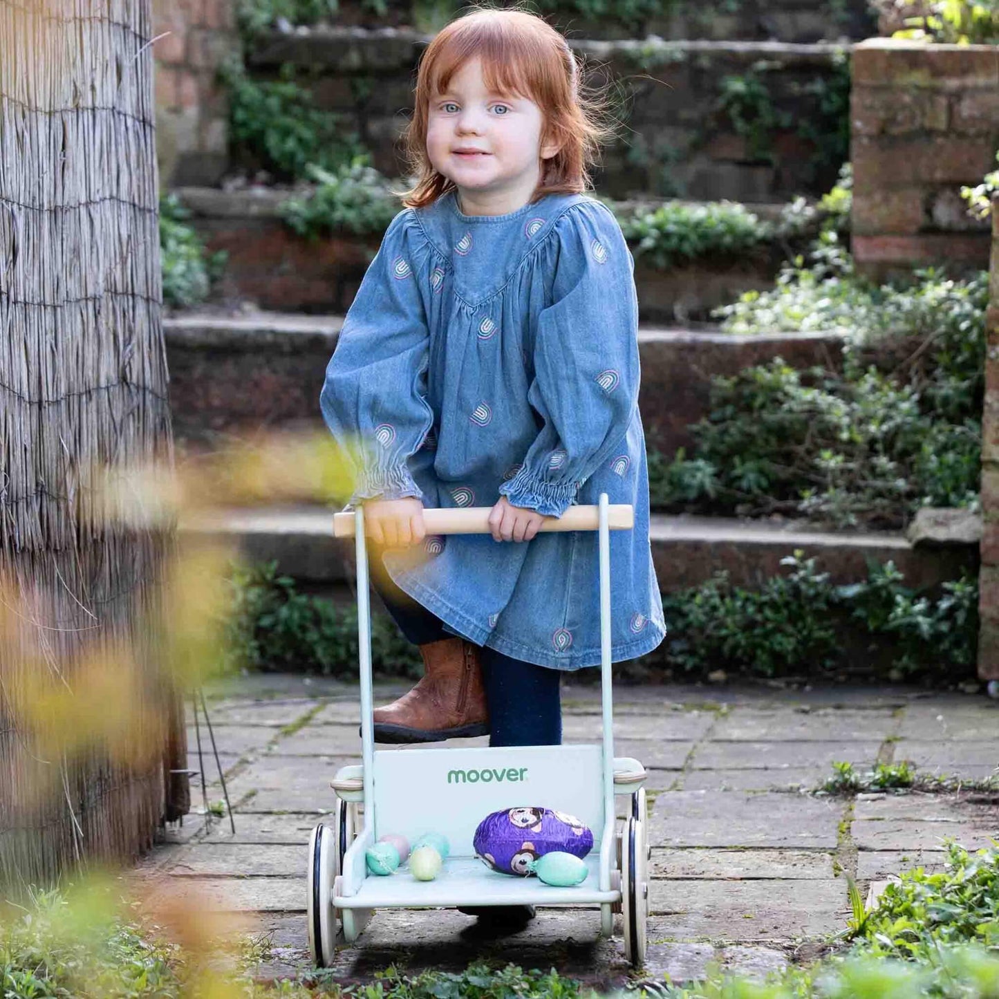 Toddler in a denim dress smiling while pushing a pale green wooden baby walker outdoors on a garden path.