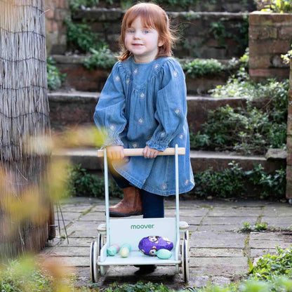 Toddler in a denim dress smiling while pushing a pale green wooden baby walker outdoors on a garden path.