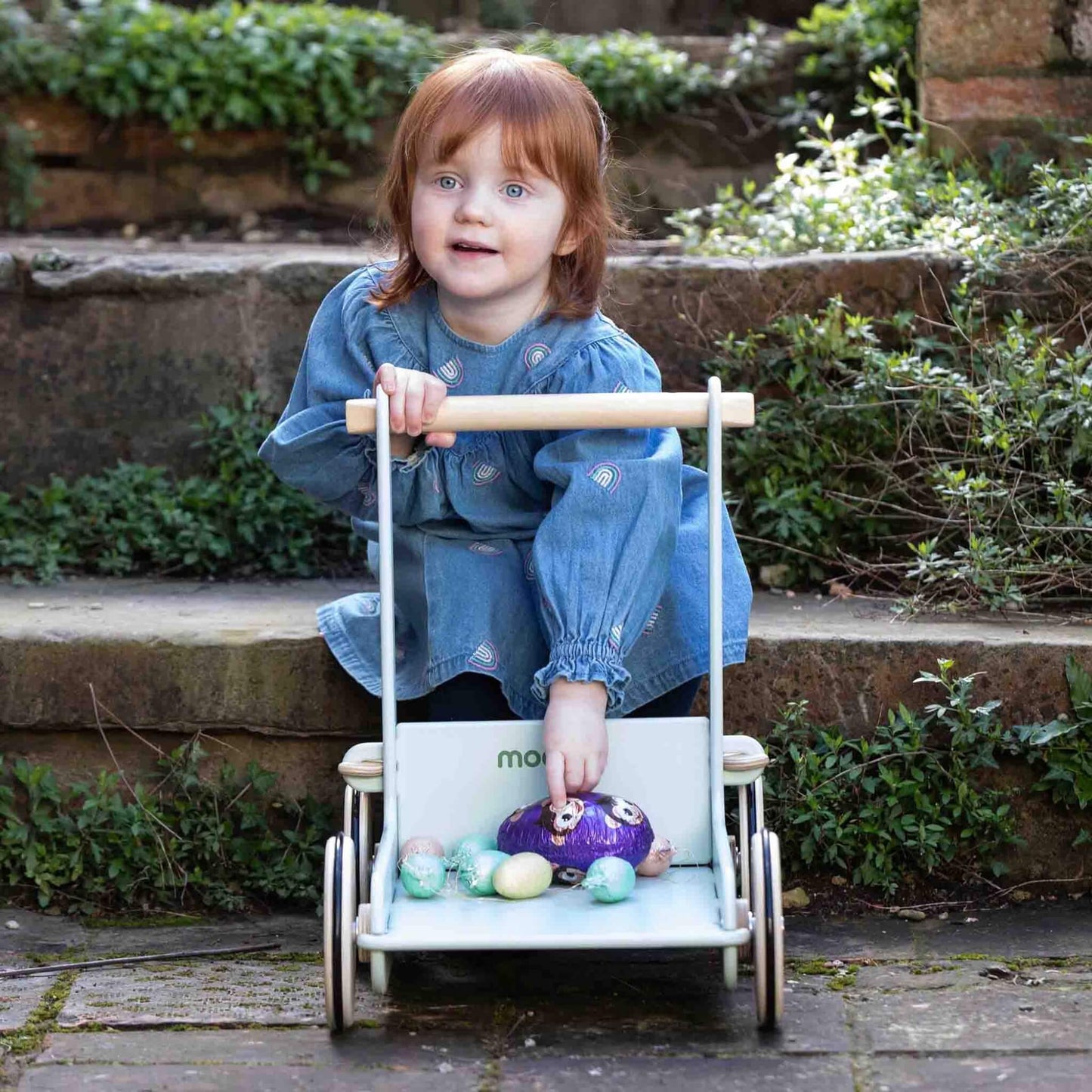 Toddler crouching to place colourful eggs inside a pale green wooden baby walker outdoors, surrounded by plants and stone steps.