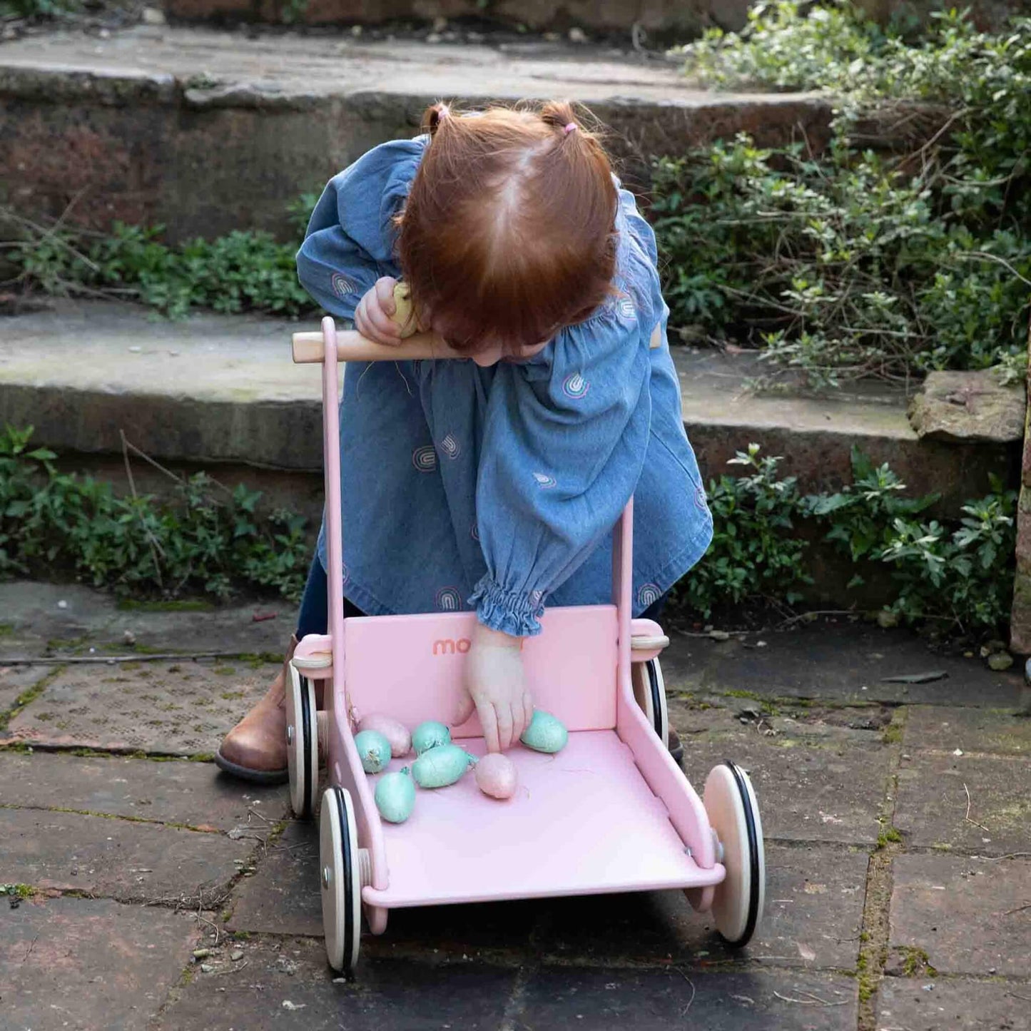 Toddler bending to place pastel toy eggs inside a pink baby walker on a garden patio.