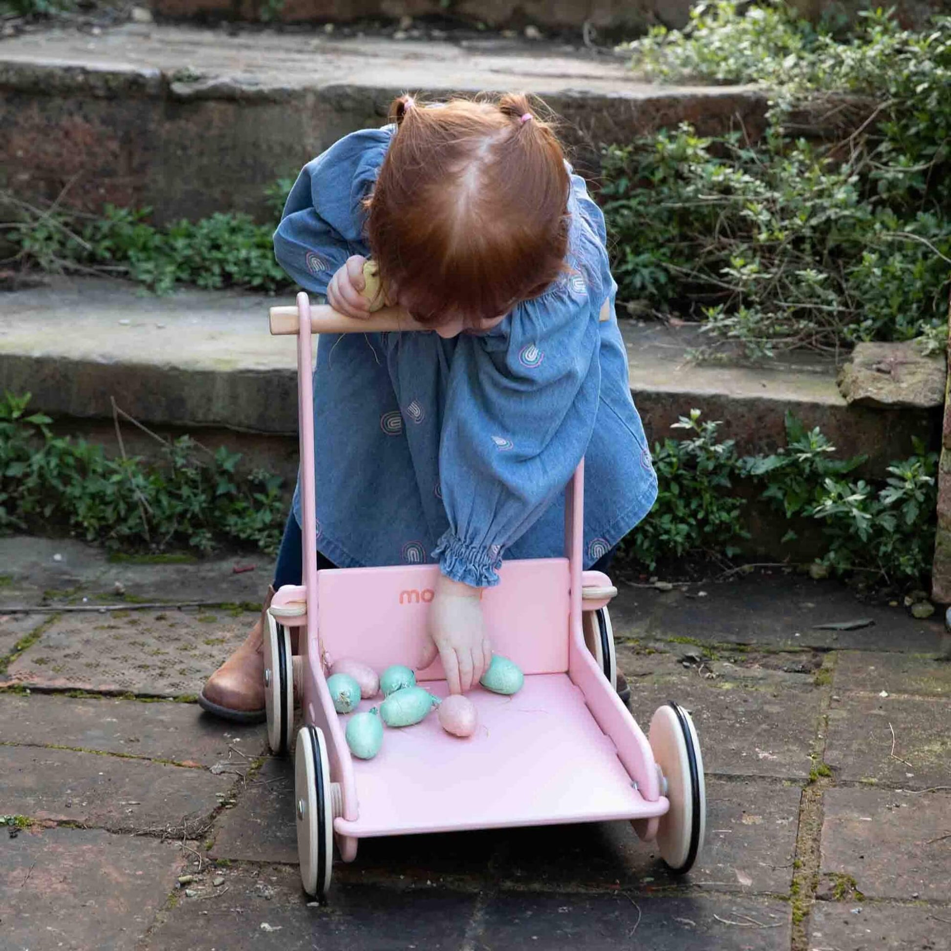 Toddler bending to place pastel toy eggs inside a pink baby walker on a garden patio.