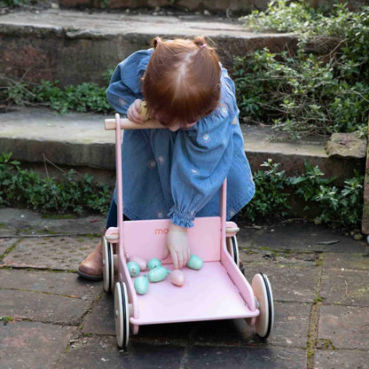 Toddler bending to place pastel toy eggs inside a pink baby walker on a garden patio.