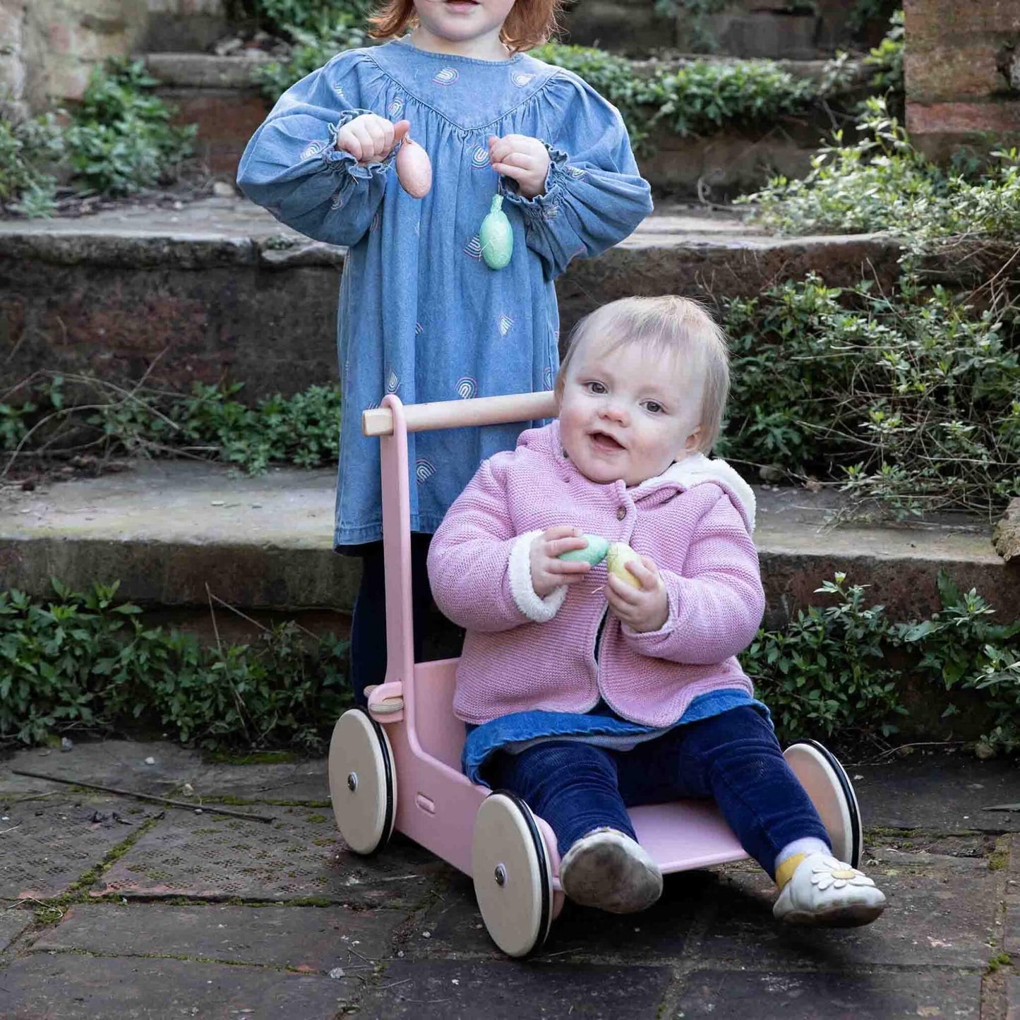 Baby sitting inside a pink baby walker while an older child stands beside holding pastel eggs outdoors.