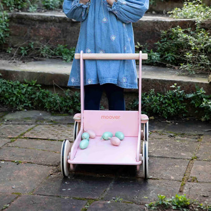 Front view of a pink baby walker filled with pastel toy eggs on a brick patio, with a child standing behind it.