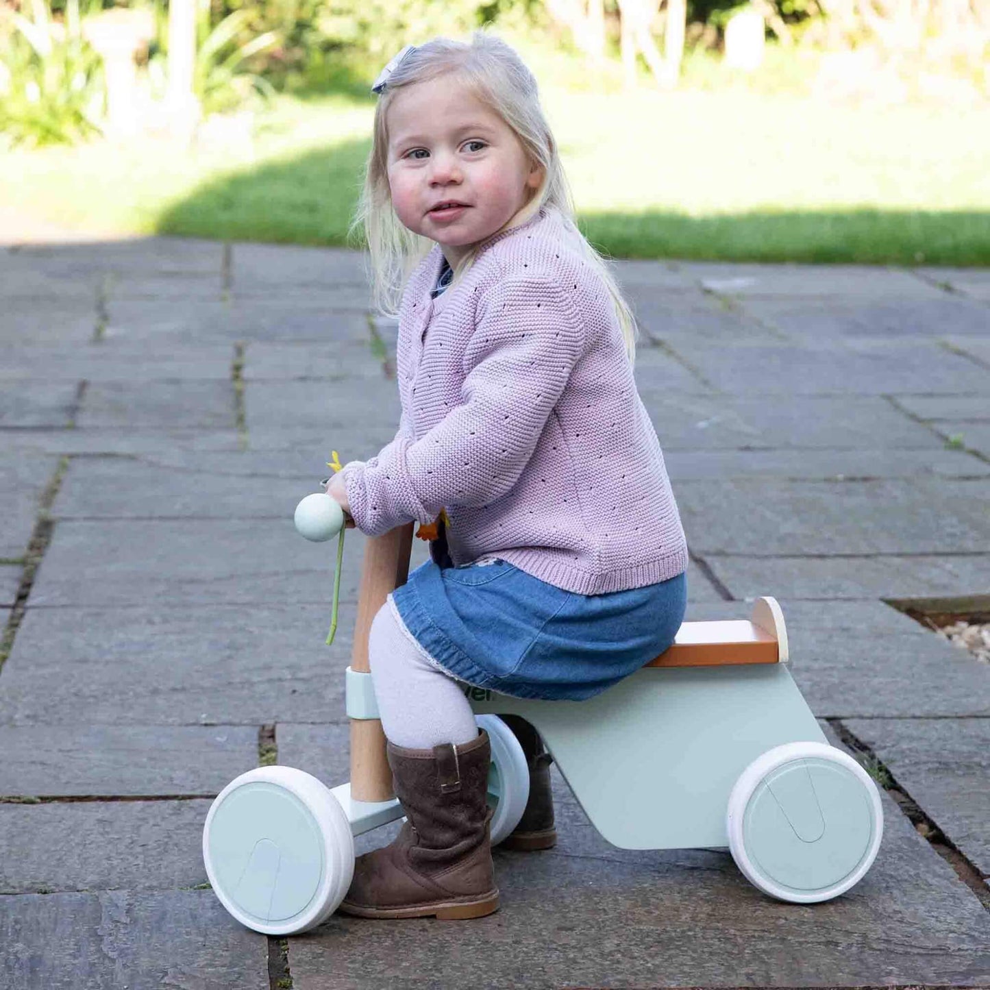 Toddler wearing denim skirt and pink jumper riding a pastel green wooden bike outdoors on stone patio.