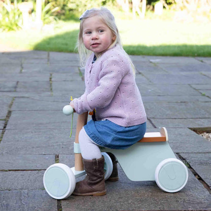 Toddler wearing denim skirt and pink jumper riding a pastel green wooden bike outdoors on stone patio.