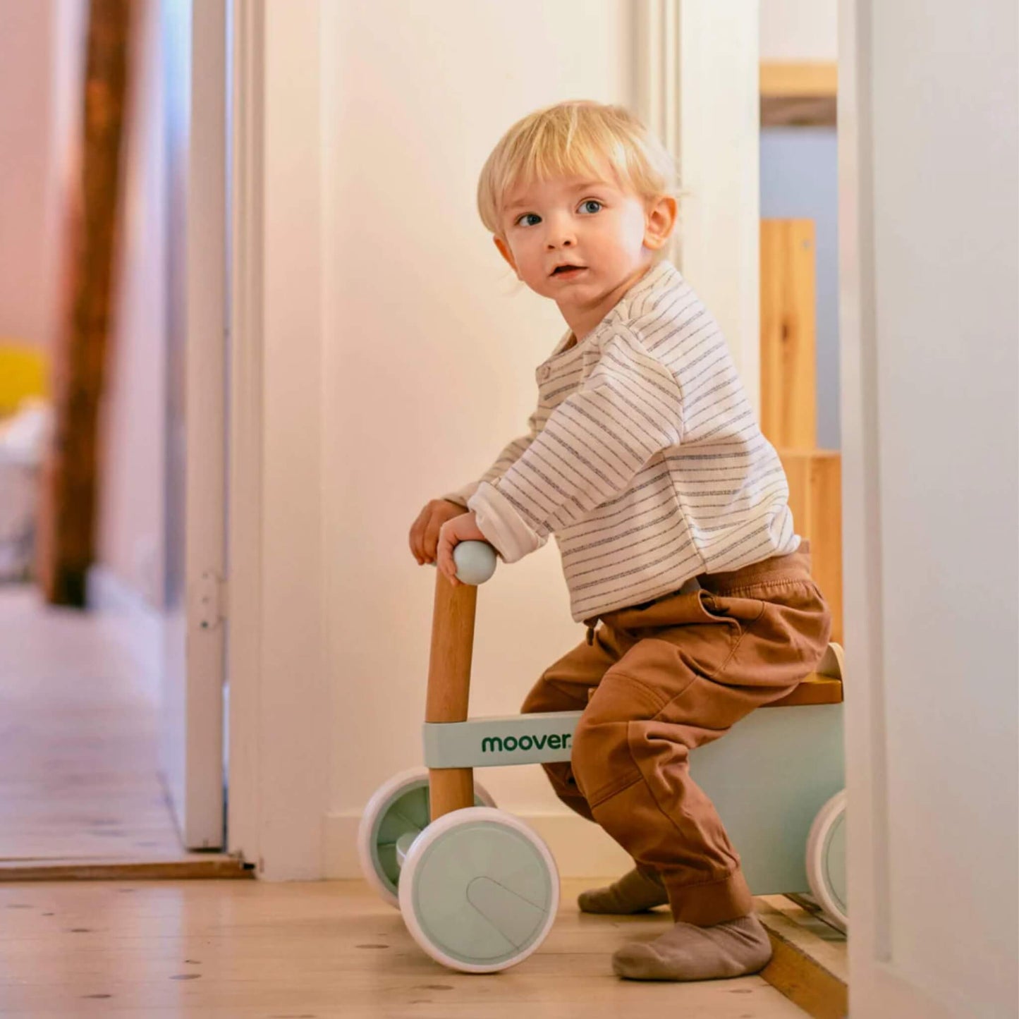 Young child in striped top and brown trousers sitting on a green ride-on bike in a softly lit hallway.