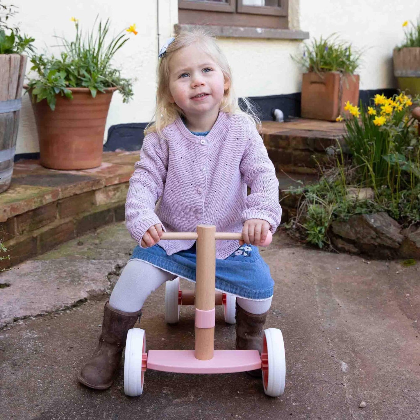 Toddler smiling while sitting on a pink wooden ride-on bike outdoors, wearing a lilac cardigan and boots near a flowered garden path.