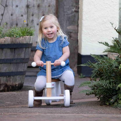 Toddler smiling while riding a white wooden balance bike outdoors, wearing a denim dress and brown boots beside potted plants.