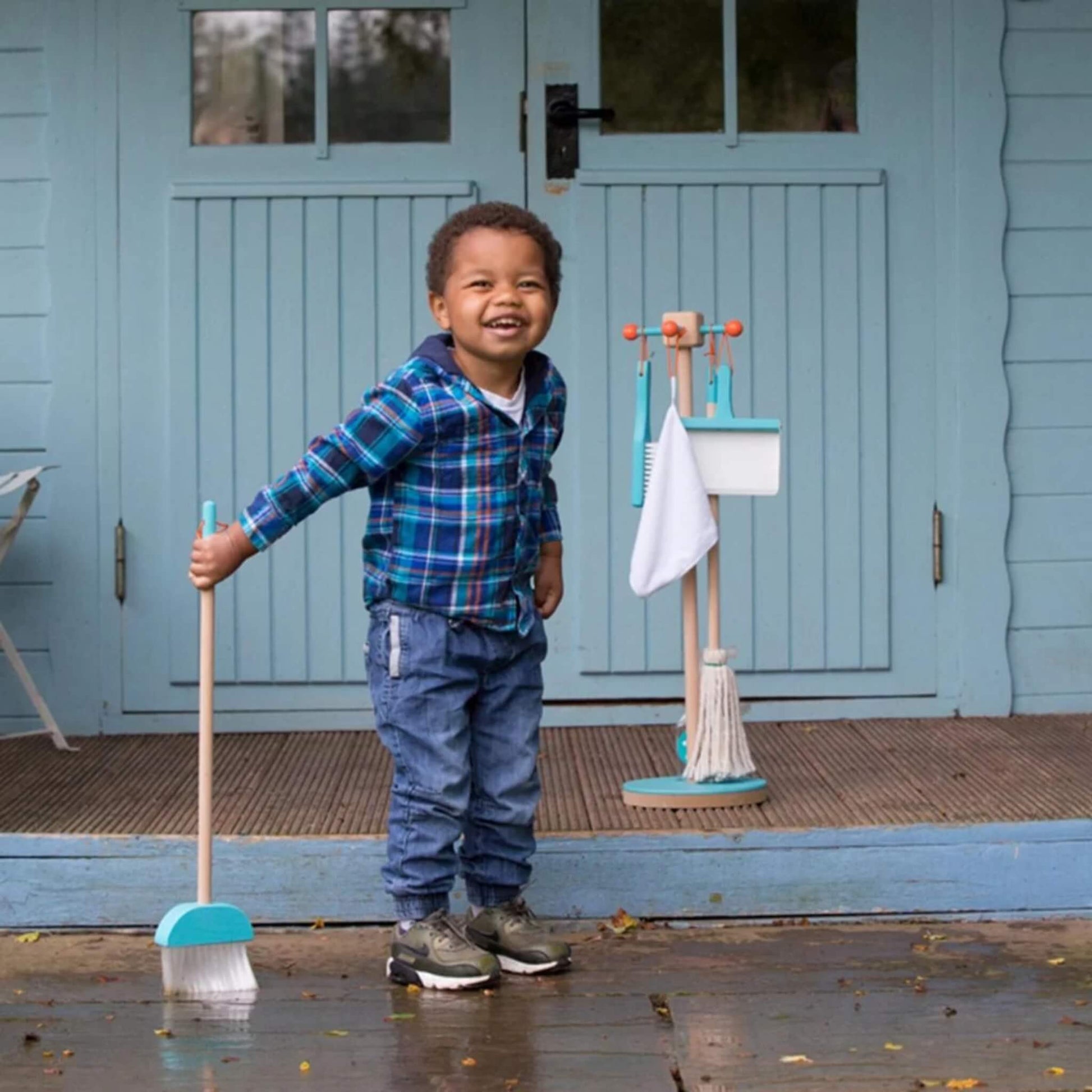 Smiling boy holding broom beside the Moover Cleaning Set in blue, standing outdoors by a playhouse.