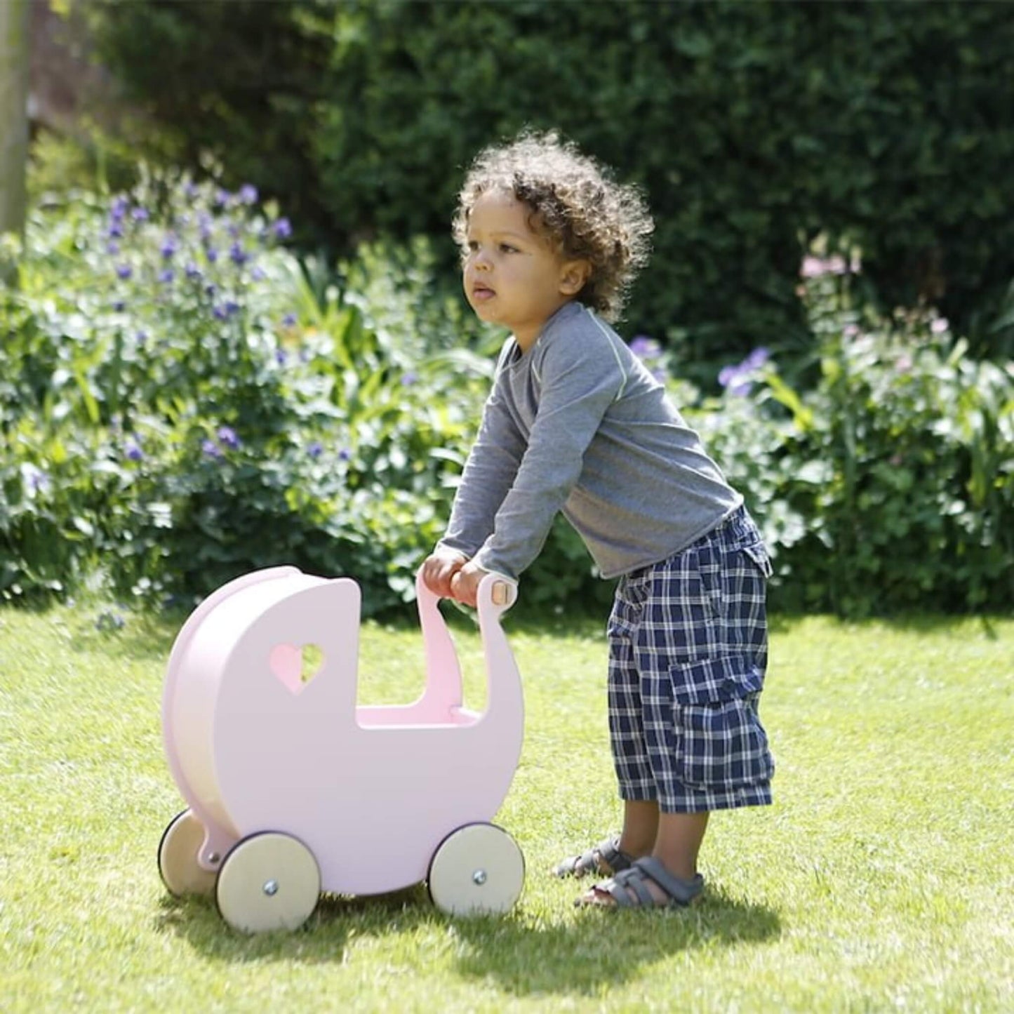 Toddler in a grey top and checked shorts pushing a pink wooden doll’s pram across a grassy garden.