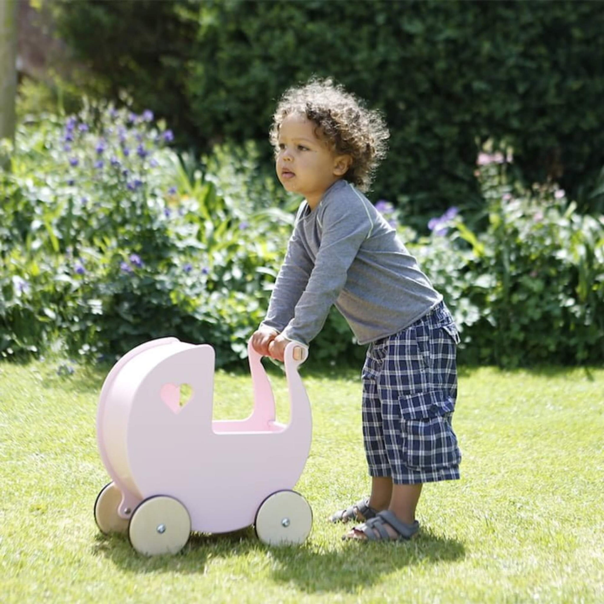 Toddler in a grey top and checked shorts pushing a pink wooden doll’s pram across a grassy garden.