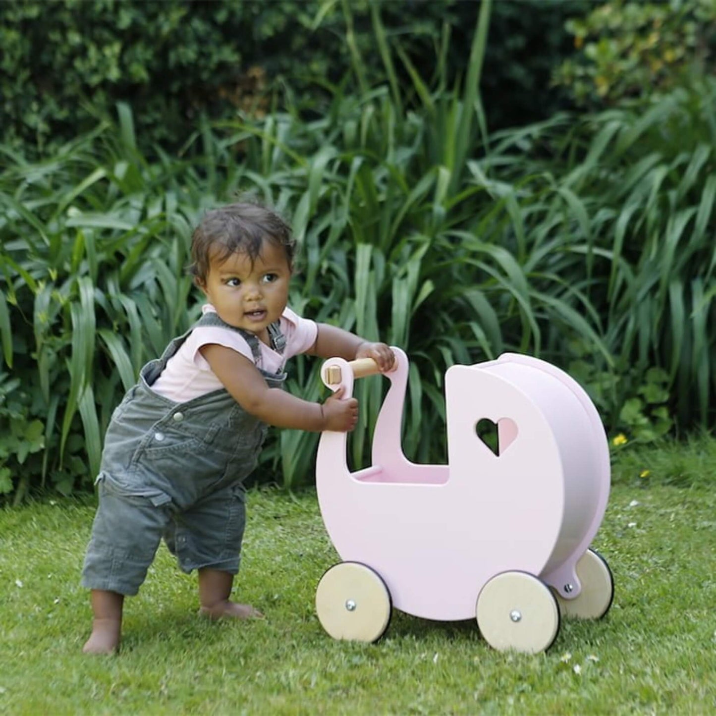 Baby wearing green dungarees and a pink top holding the handle of a pink wooden doll’s pram in a garden setting.