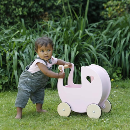 Baby wearing green dungarees and a pink top holding the handle of a pink wooden doll’s pram in a garden setting.